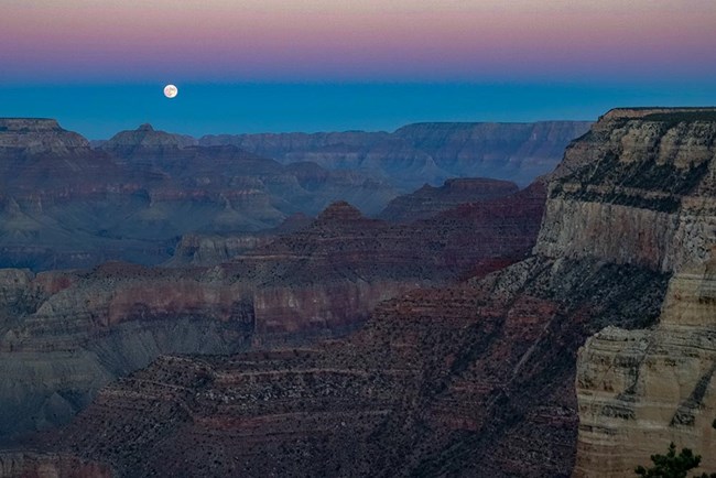 Full Moon seen rising above Vishnu Temple, just after the sun set on the horizon. This wider view of the mile deep canyon landscape is from Powell Point on the South Rim. Cliffs, ridgelines and buttes are seen under the pinkish glow