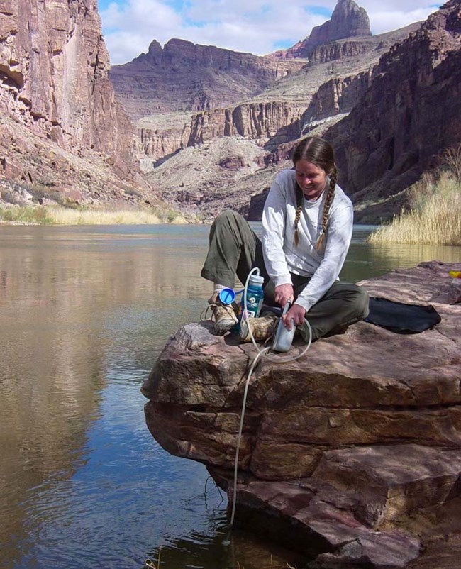a woman sitting on a rock ledge several feet above a river is filtering water through a tube hanging down from the ledge into the water. Cliffs and peaks visible in the distance.