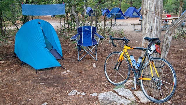 a yellow bicycle next to a tree and a blue tent, with a row of blue tents in the forested background.