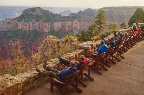 about one dozen people sitting in wooden deck chairs and looking over the stone patio wall at Grand Canyon during sunset.