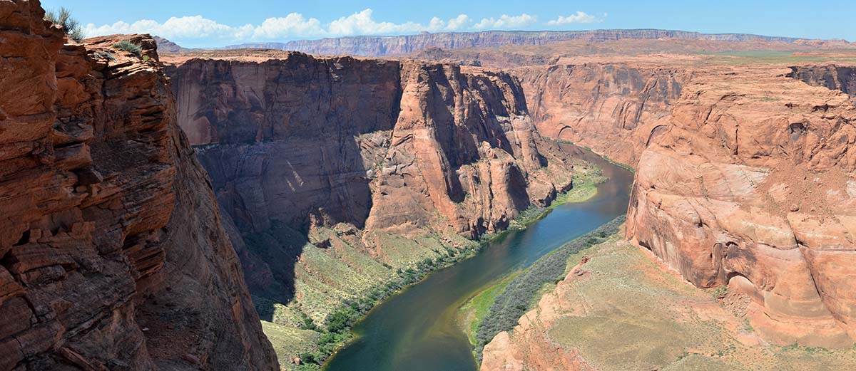 Looking down from the top of a sheer sandstone cliff at a green river at the bottom. Foliage growing along the shoreline.