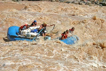 A boatman rowing a blue inflatable raft through a brown, churning Colorado River rapid. Four passengers are in the boat, two in front, and two in back.