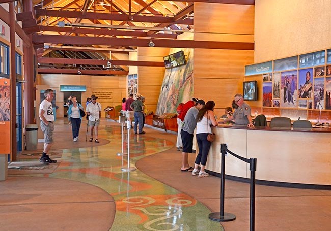 a large room with high ceilings and exhibits and maps on the walls. standing behind a curved desk, a park ranger is answering visitor questions.