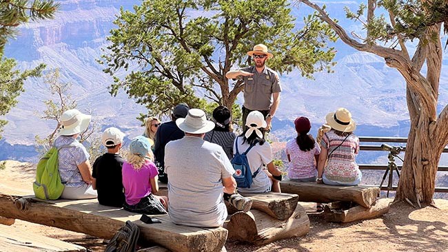 a park ranger standing in an outdoor amphitheater is giving a talk to a group of park visitors. He is using hand gestures to explain geologic processes. Grand Canyon is behind them.