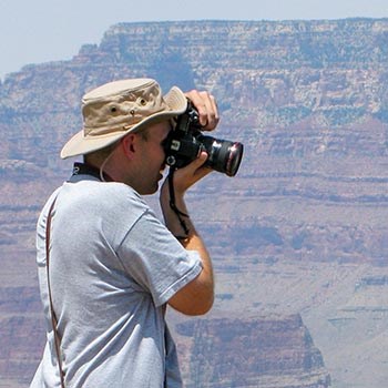 A person wearing a floppy sun hat is photographing outdoors with canyon walls in the background.