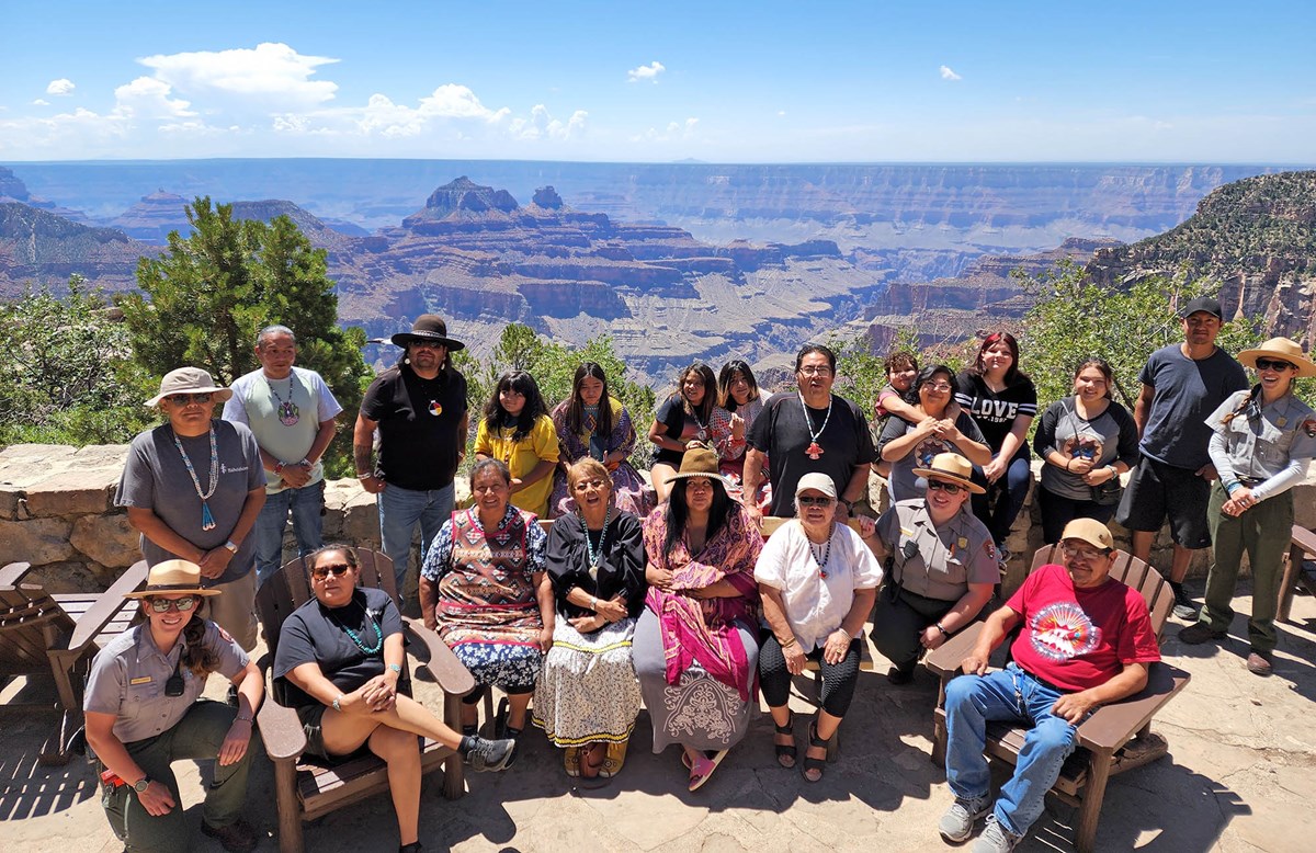 22 people arranged in two rows are posing for a group photo by a stone wall overlooking several peaks that rise from the floor of a vast canyon.