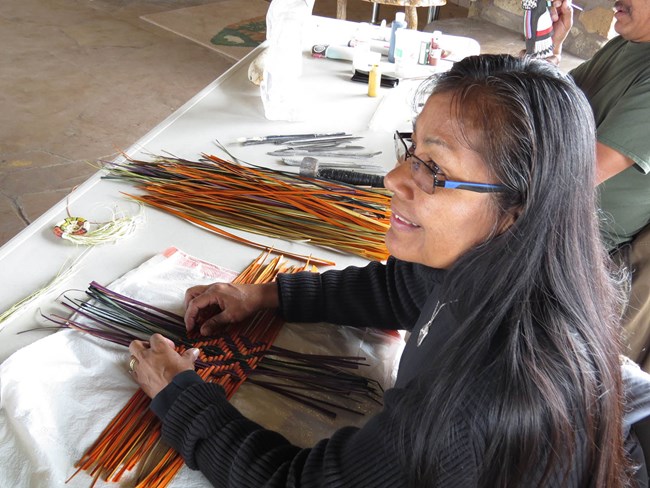 a Hopi woman with long black hair is weaving a basket with orange, yellow and black strands of yucca leaves.