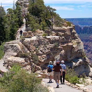 Several day hikers on the paved footpath with ascending and descending slopes to Bright Angel Point. A view of peaks within Grand Canyon are visible in the distance on the right.