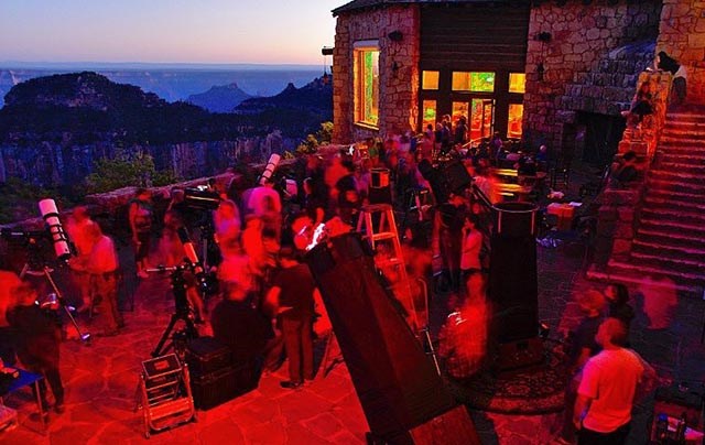 Twilight; lluminated by red light, a group of people are looking through telescopes on the veranda of a rustic lodge.