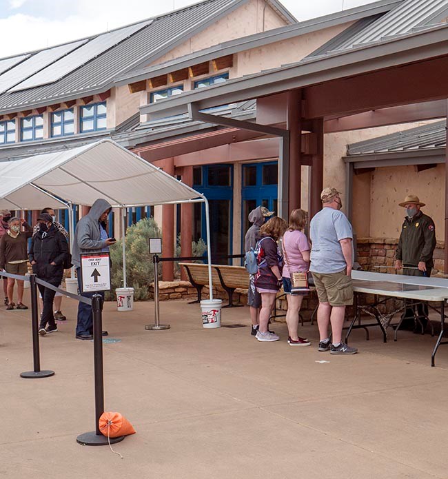 On the left, several people waiting in line under tent canopy to ask questions of a park ranger standing behind an outdoor information table with maps.
