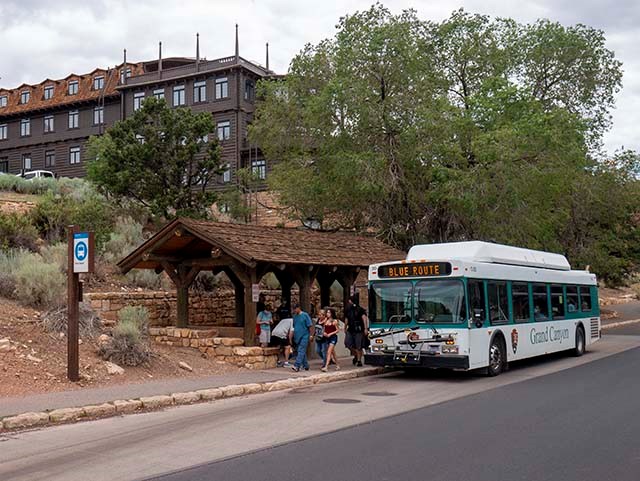 a white and green bus is unloading passengers by a rustic open bus stop with a shake roof. in the background, a four story hotel.