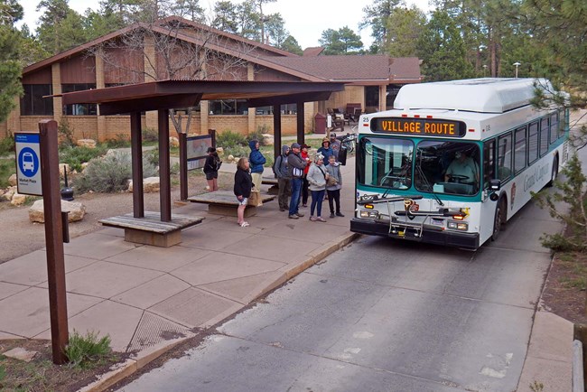 from under the roof of an open air bus shelter, several people are about to board a white and green bus