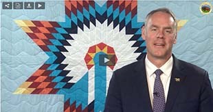 Secretary of the Interior Zinke wearing a blue suit and tie, is standing in front of a quilt decorated with an American Indian design.