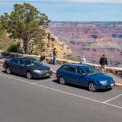 two cars parked by the side of the road at a scenic overlook. Behind a stone guard wall, four people are viewing a colorful canyon landscape of peaks and cliffs.