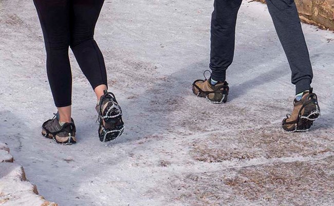 two people wearing metal over the shoe traction devices are walking on an icy footpath.