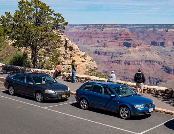 Two cars are parked at a pullout for a scenic overlook. Four sightseers are standing behind a stone guard wall and viewing a vast and colorful canyon landscape in the distance.