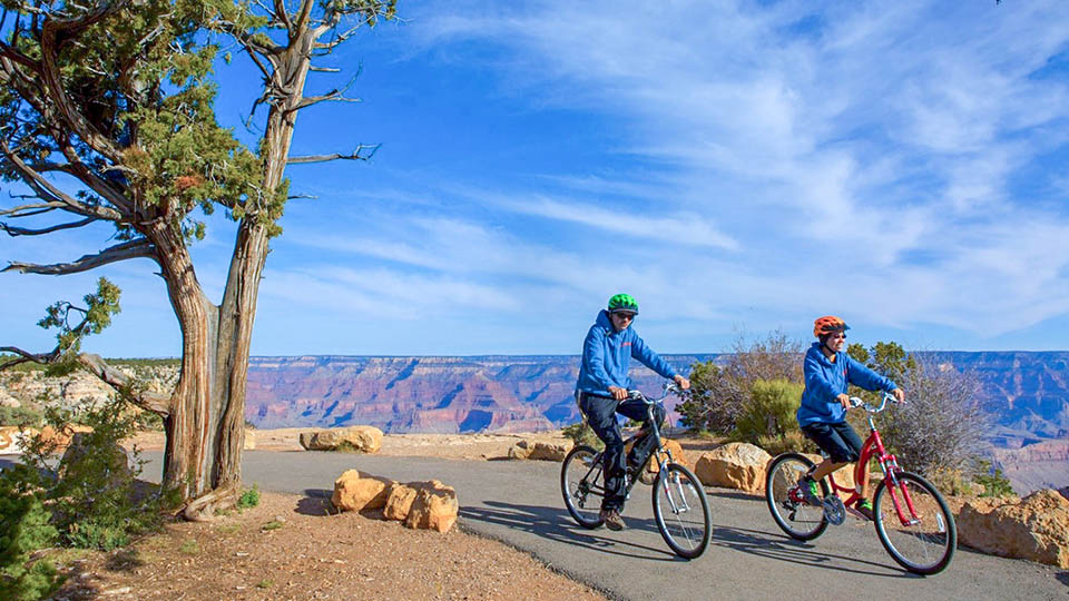 Image: 2 bicyclists riding along a paved greenway path along the edge of a colorful canyon landscape