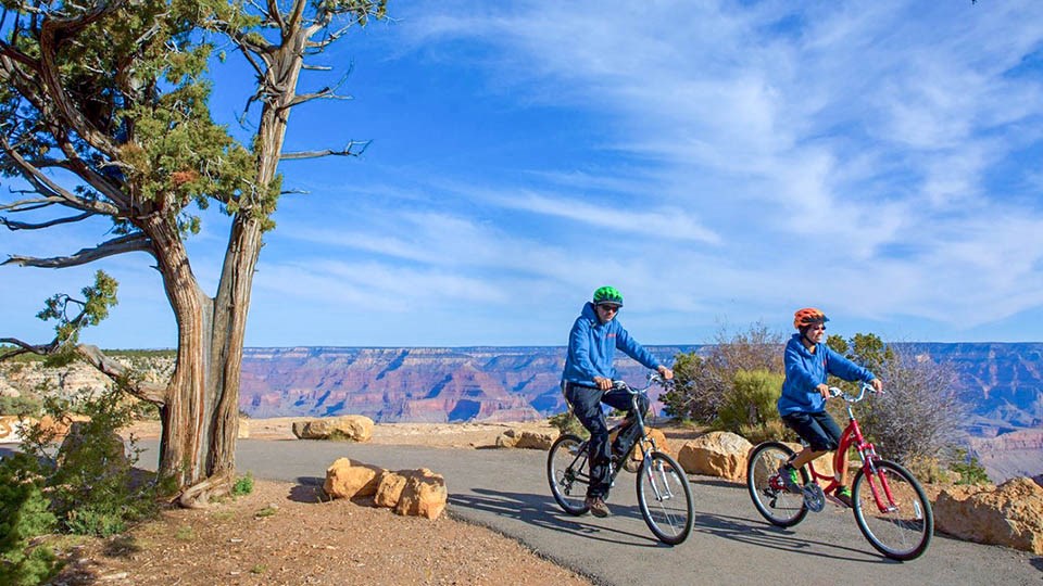 Image: 2 bicyclists riding along a paved greenway path along the edge of a colorful canyon landscape
