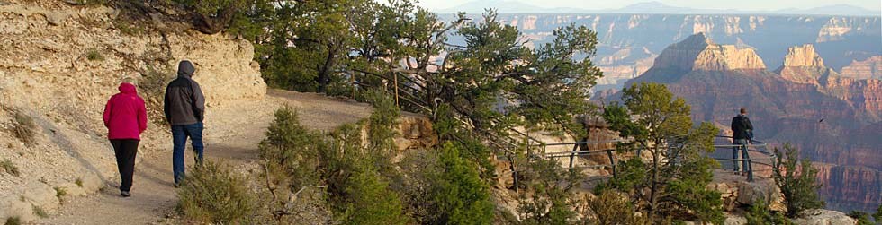 two people walking on a trail. a scenic overlook is on their right with Grand Canyon in the distance