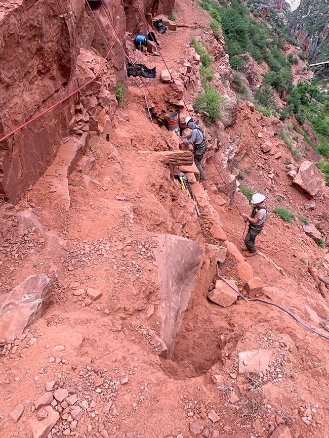 On the side of a cliff, two trail crew workers, wearing helmets, are repairing a section of damaged trail. A handline traverses an area that is only a narrow ledge.