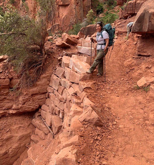 a woman holding trekking poles and wearing a backpack is standing above a stone wall created to replace a section of trail that was destroyed by a landslide.