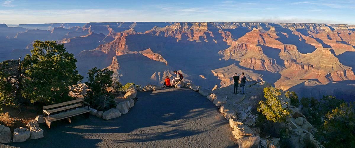 four people at a scenic overlook are experiencing a vast mile deep canyon landscape filled with colorful cliffs and peaks. Illuminated areas are standing out from deep shadows.