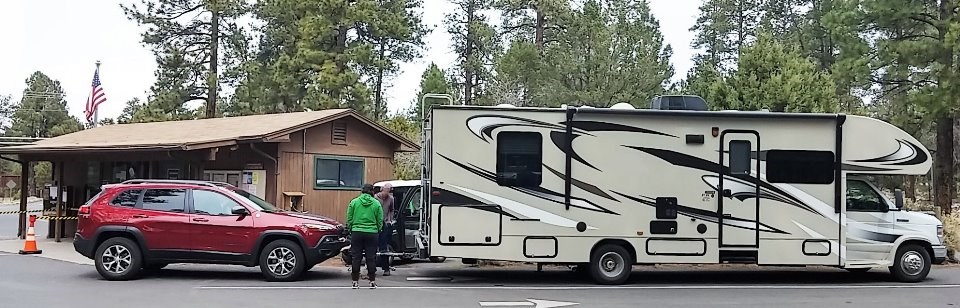 a large white RV is stationary with a red car in tow.. Two people are disconnecting the car from the RV. This is in front of a brown campground office.