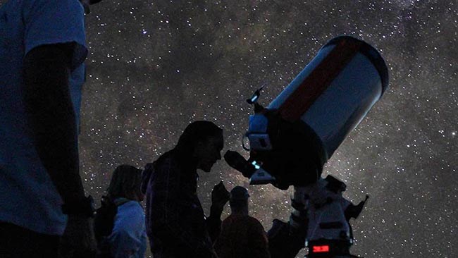 a small group of people taking turns looking through a large telescope beneath a sky full of stars.