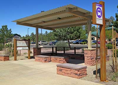 Typical Tusayan shuttle bus stop/shelter have red stone benches beneath a olive color corrugated metal roof. There is also a route map exhibit and an elevated sign on a pole that identifies each stop.