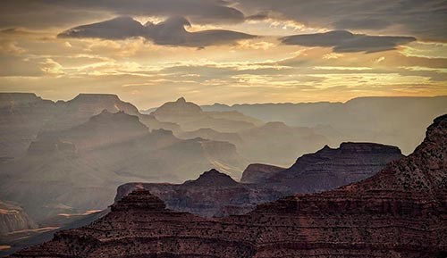 During sunrise, sunbeams visible through silhouetted peaks and ridgelines within a vast canyon landscape. A variety of clouds in different shapes fill the sky.