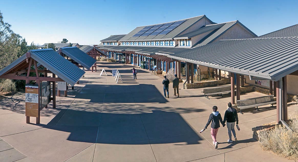 view down and across a concrete plaza with a long narrow building with a gabled roof on the right. several people are walking towards the building.