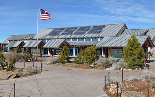 Within a paved plaza is a long, narrow building with solar panels on a gabled gray roof. Five exhibit kiosks are positioned in front of the building and the American Flag is posted in front of the building.