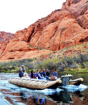A large motorized raft holding eight people on board is traveling down a river with orange sandstone cliffs in the background, with vegetation along the shoreline.