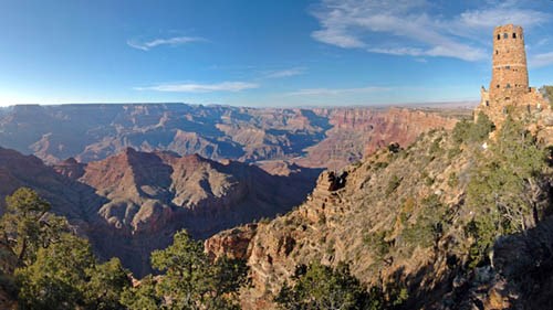 Looking across the vast expanse of Grand Canyon landscape towards a round, stone Watchtower on the far right, perched on the edge of a cliff.