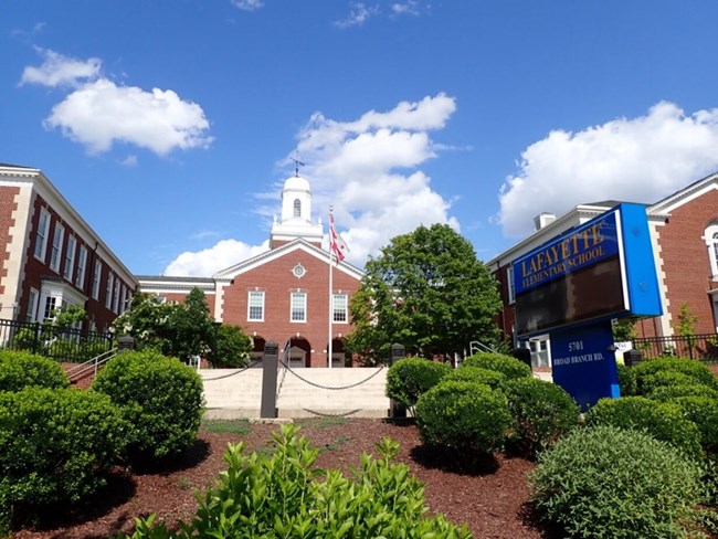 A photo of a red and white brick school house, on a sunny day, with an American Flag and a blue school sign in the foreground