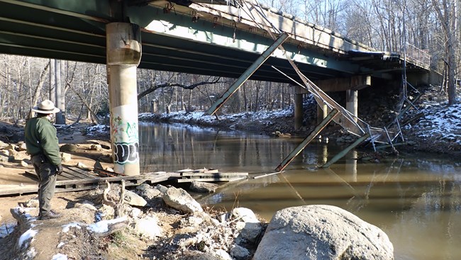 A NPS Park Ranger stands on a dirt and rocky trail with wooden sidewalk debris across the trail next to a creek with additional sidewalk debris collapsed and hanging from a bridge above.