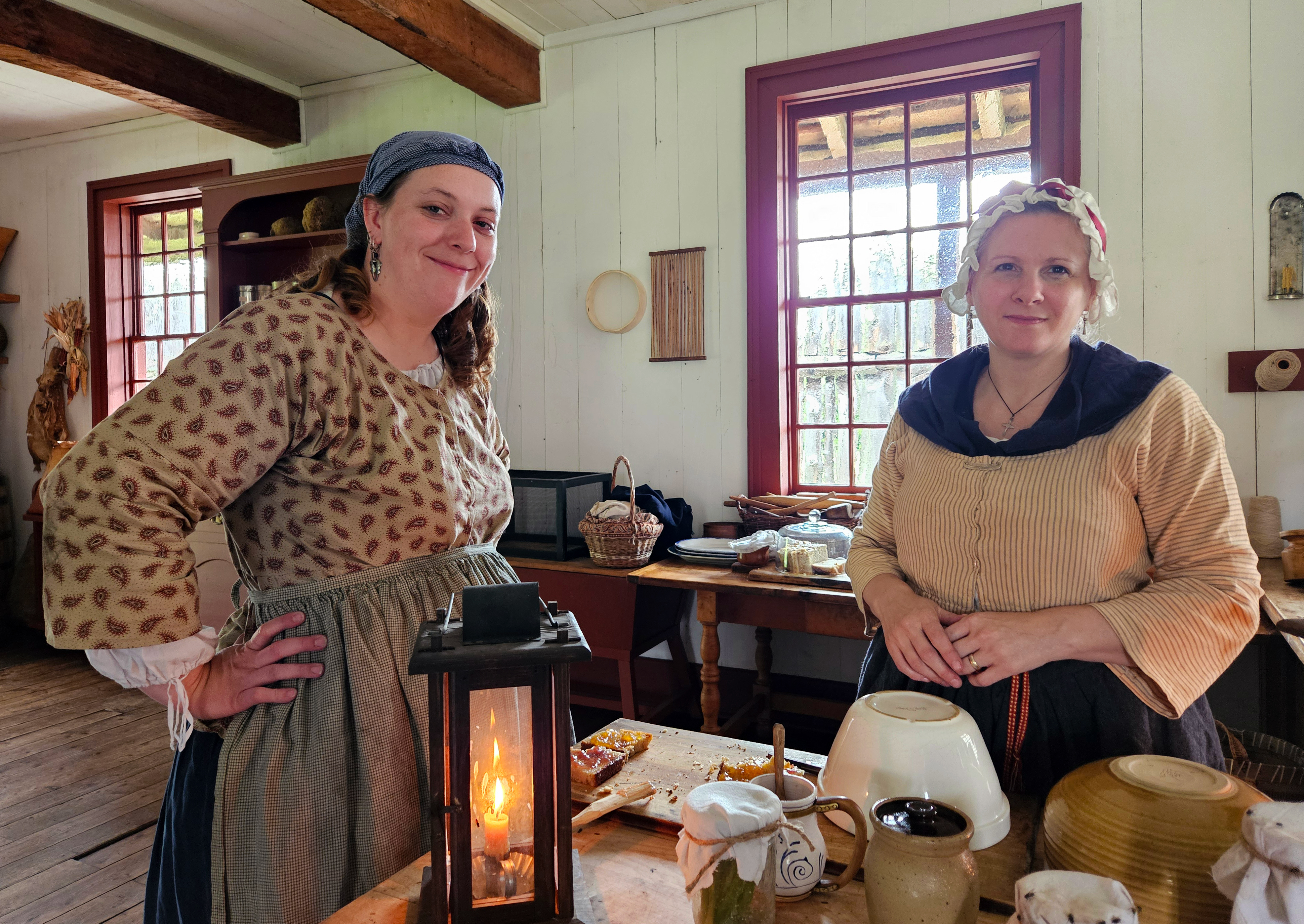 Two people in historic clothing stand around a table with a lantern and several pottery containers.