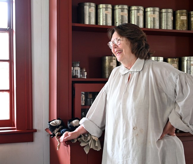 Volunteer in historic white shirt leaning on a reddish brown kitchen shelf unit.