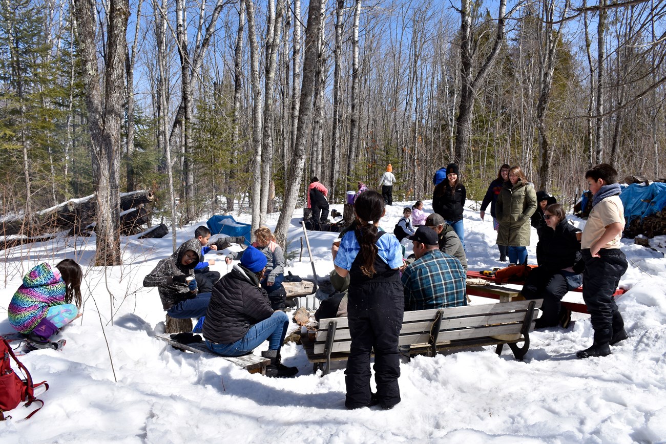 People wearing winter clothing gathered around a fire in a snowy forest.