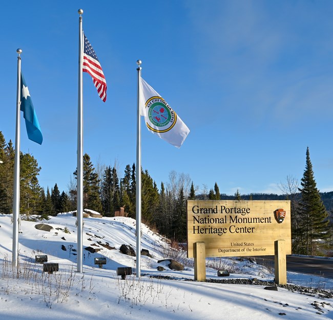 Three flags: Minnesota state, US, Grand Portage Band, stand next to a wooden NPS style sign for Grand Portage National Monument Heritage Center with the National Park Service arrowhead logo.