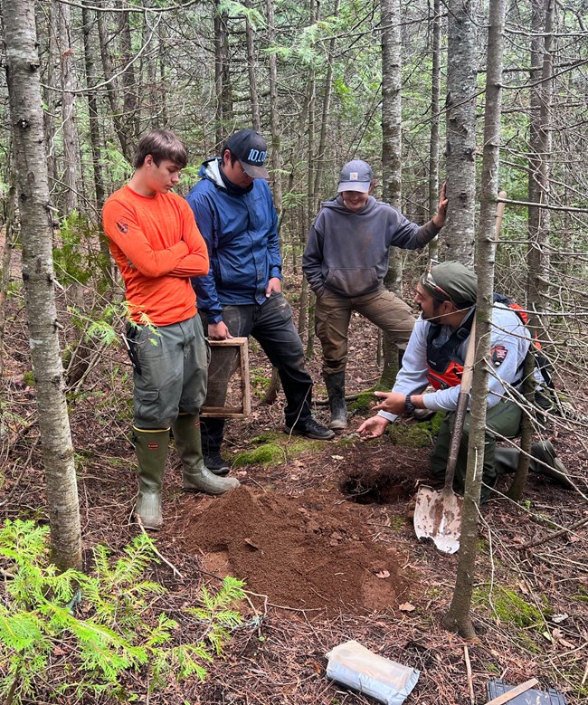 Three people standing and one kneeling in a forest around some loose dirt.