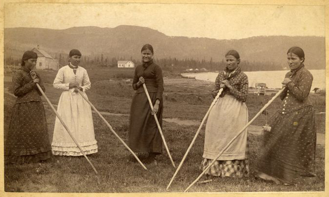 Black and white photo of five women in historic clothing holding long sticks with one end on the ground in a half circle.