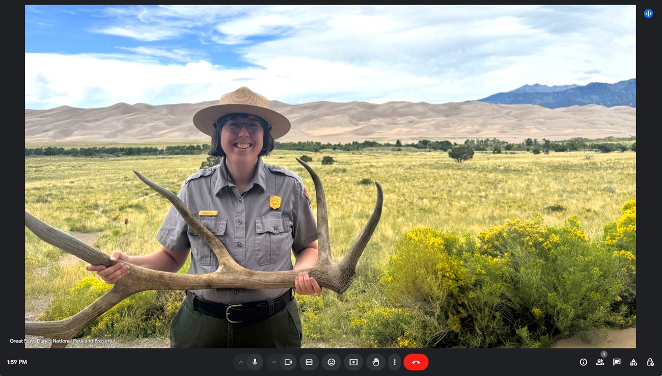 A park ranger holds an elk antler with grasslands, dunes, and mountains in the background