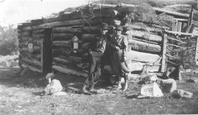 A historical black and white photo of a log structure with a man and woman standing beside it