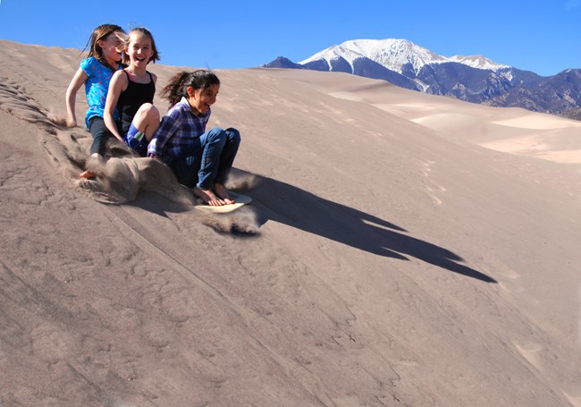 Three girls ride on a sled down a dune, with a snow-capped mountain behind them
