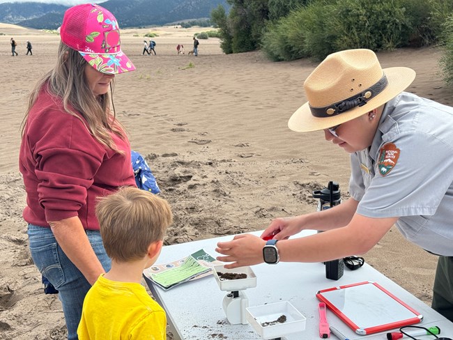 A Park ranger in green and grey uniform with straw ranger hat leans over a table to help a child with a magnetic sand activity