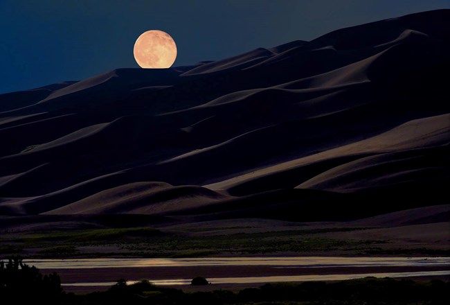 The full moon hovers over the dunes at night as Medano Creek reflects its light in the foreground