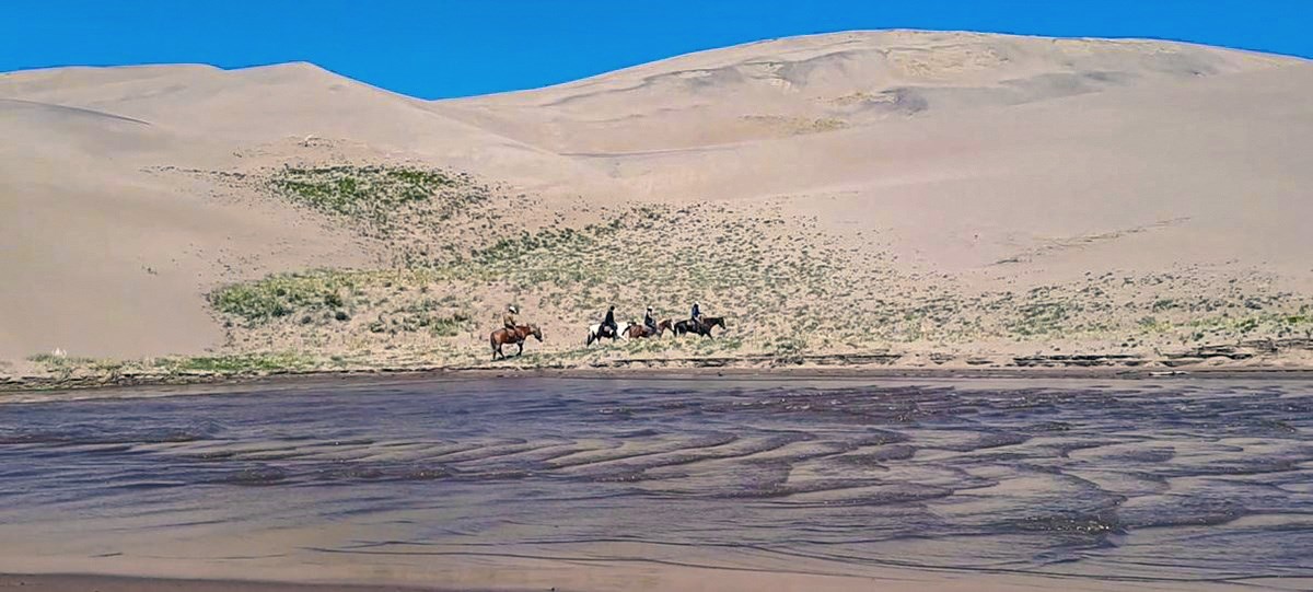 Four horseback riders on the dunes above a shallow stream