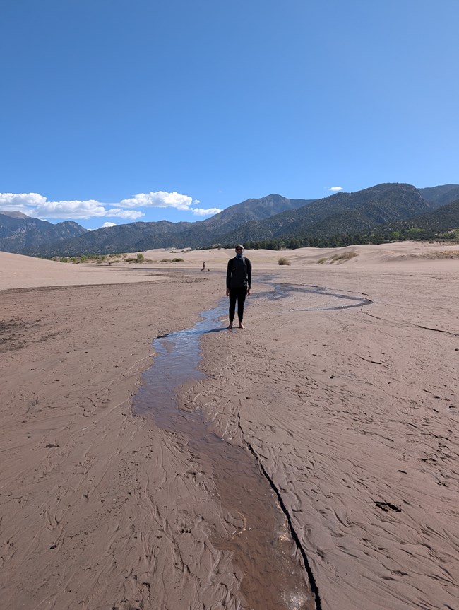 A park visitor stands at the end of Medano Creek. The creek is a foot wide and under an inch in depth as it flows over sand. Tall dunes and mountains rise in the background.