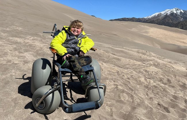 A child sits in a dunes wheelchair on a dune, with a mountain in the background.